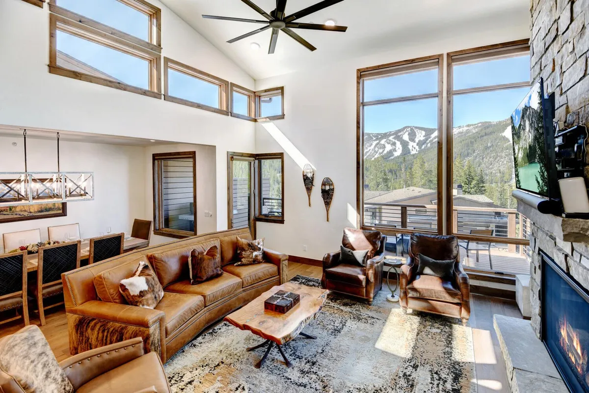 A spacious, sunlit living area featuring large windows that frame breathtaking mountain scenery. The room is furnished with a comfortable leather sofa, a rustic coffee table, and artistic wall decor, creating an inviting space ideal for family relaxation and entertaining guests.