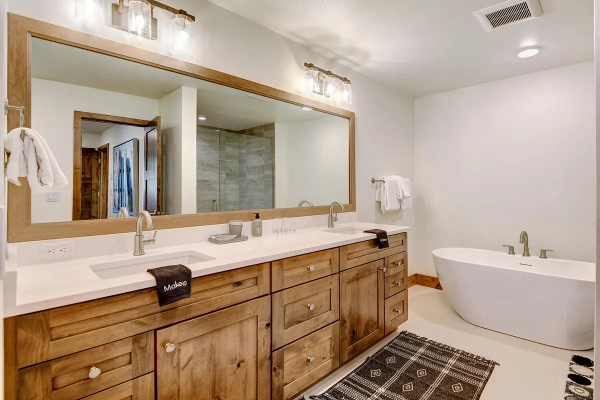 A spa-inspired bathroom featuring a double sink vanity, a freestanding soaking tub, a large mirror, wood cabinetry, and a decorative rug. The calming and inviting atmosphere reflects a high-end, indulgent experience.
