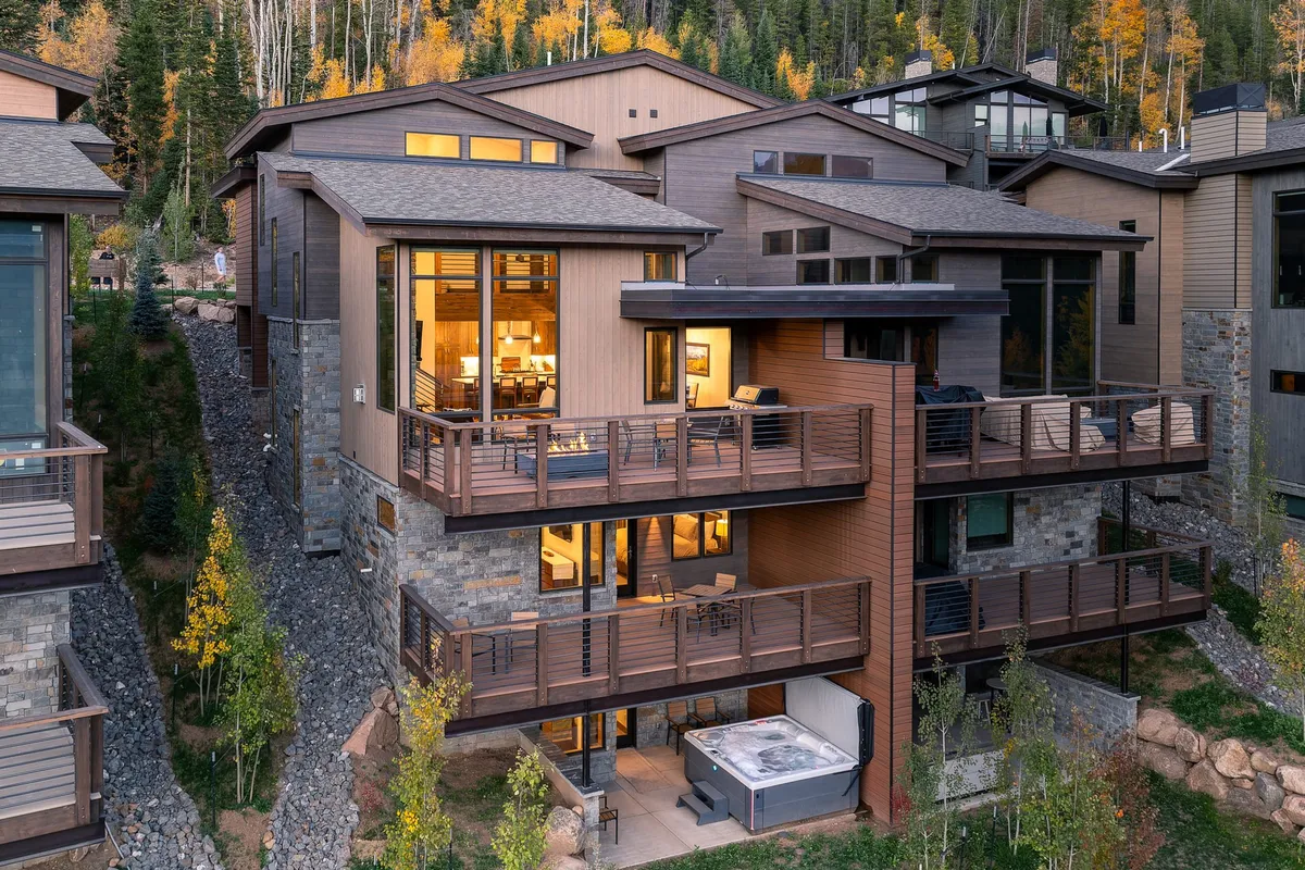 The image showcases a modern, luxurious mountain villa with expansive decks, a private outdoor hot tub, and panoramic views of natural Colorado scenery. The scene evokes relaxation and indulgence—guests can be seen enjoying the hot tub, socializing on the spacious terrace, and soaking in the stunning sunset over the mountains.