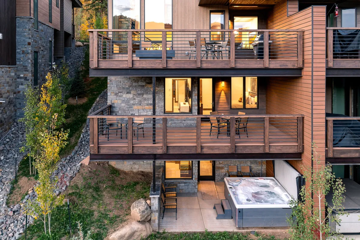 This exterior image highlights the villa's balconies, a bubbling hot tub with mountain views, and large windows that hint at the spacious interiors. The scene conveys a sense of luxury and relaxation, with seating areas perfect for group gatherings or unwinding after adventurous activities.
