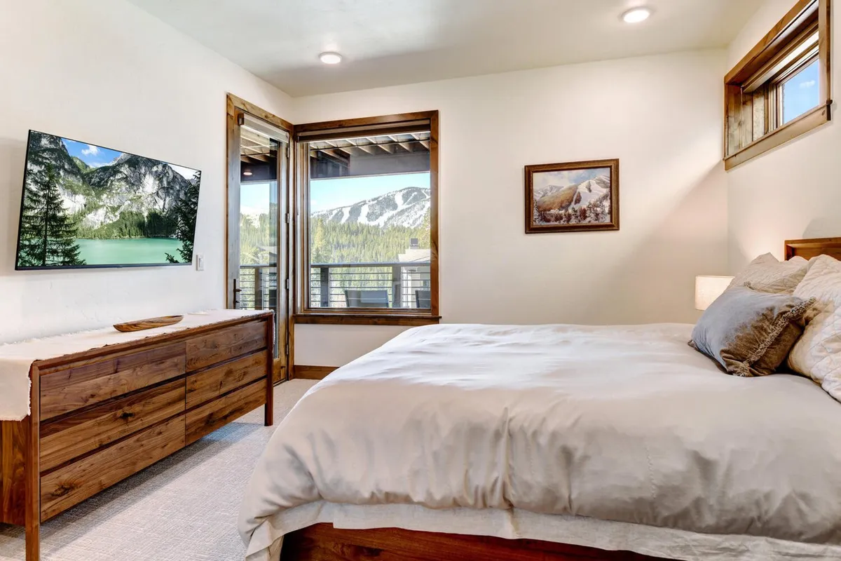 A tranquil and comfortable guest bedroom with a plush bed, a television, a wooden dresser, framed artwork, and a window offering scenic views. The space feels welcoming and peaceful, ideal for relaxation and privacy.