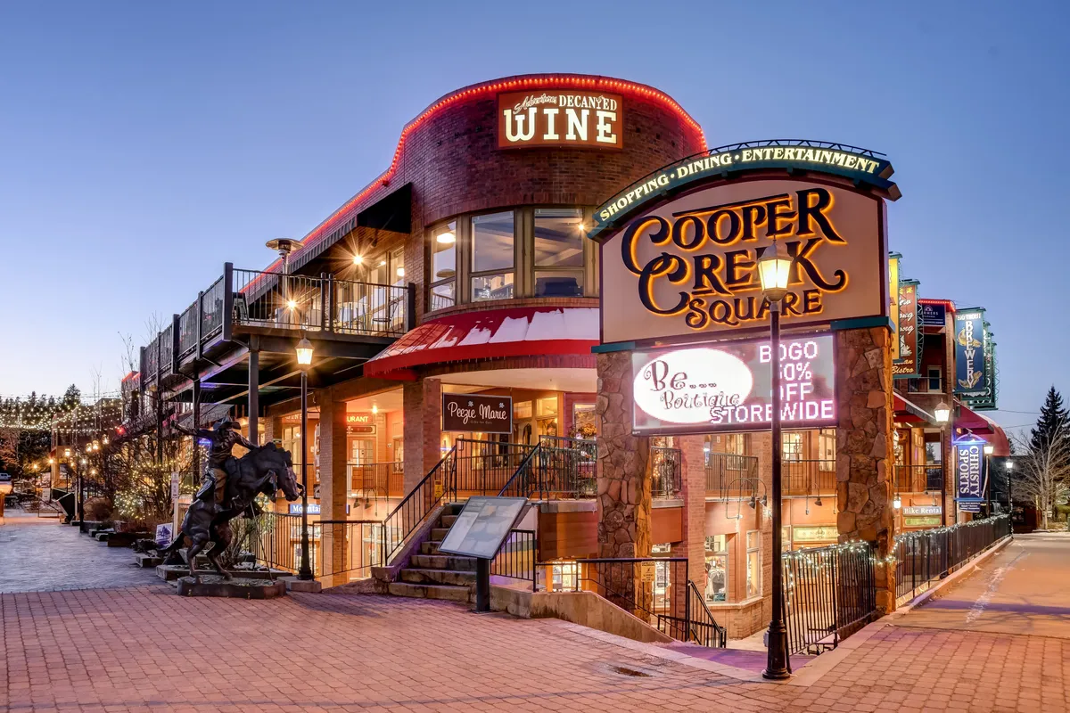 The vibrant Cooper Creek Square sign, adjacent Decadent Wine sign, outdoor seating, and horse sculpture capture the lively atmosphere of downtown. The setting is festive and social, hinting at the rich local dining and entertainment options accessible from the villa.
