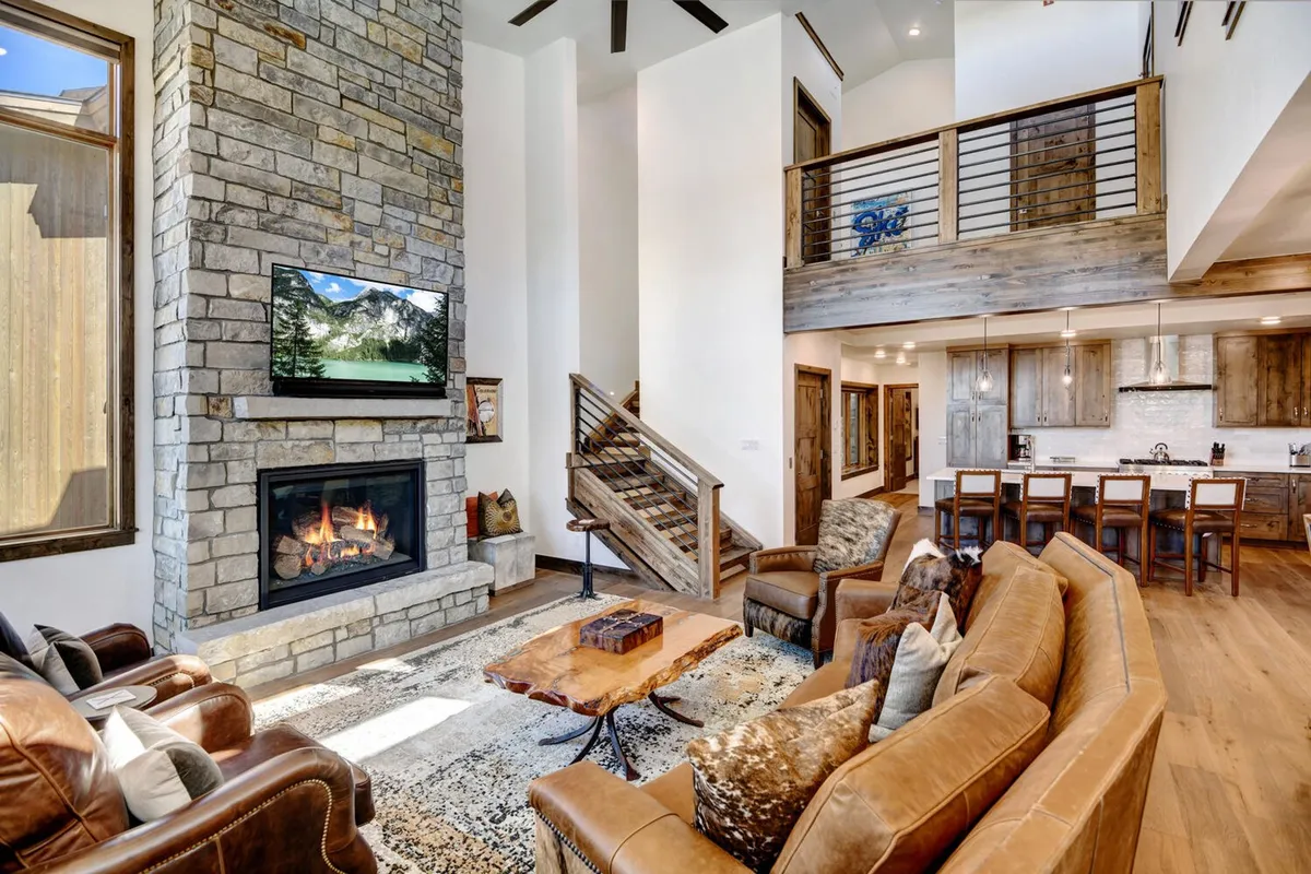 A spacious lower-level den featuring a large sectional sofa facing a wall-mounted TV above a rustic stone fireplace. The room exudes warmth with its plush seating, inviting gathering spaces, and a wooden coffee table, creating an ideal spot for movie nights, watching the big game, or simply relaxing by the fire.