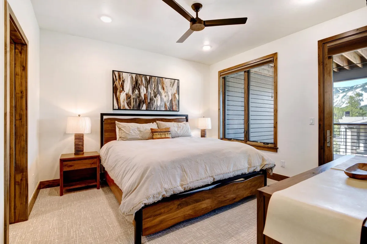 A serene king-sized bedroom featuring a large bed with decorative pillows, nightstands with lamps on each side, tasteful wall art, and a generously sized window that allows natural light to brighten the space. The soothing palette and upscale finishes create a tranquil, luxurious atmosphere.