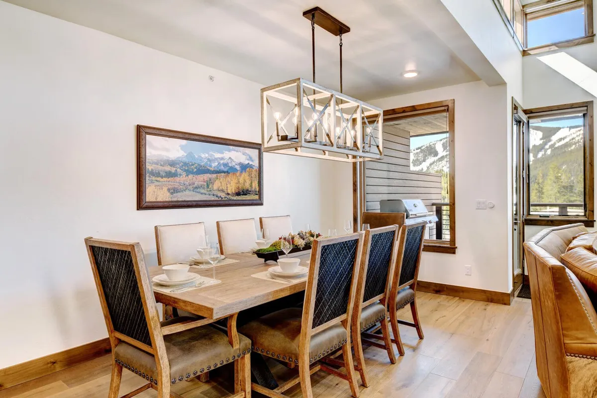 This scene centers on a large dining table set beneath a chandelier, with a backdrop of a mountain view painting and plenty of natural light from expansive windows. The table is suited for seating eight, ideal for group gatherings and celebratory meals.