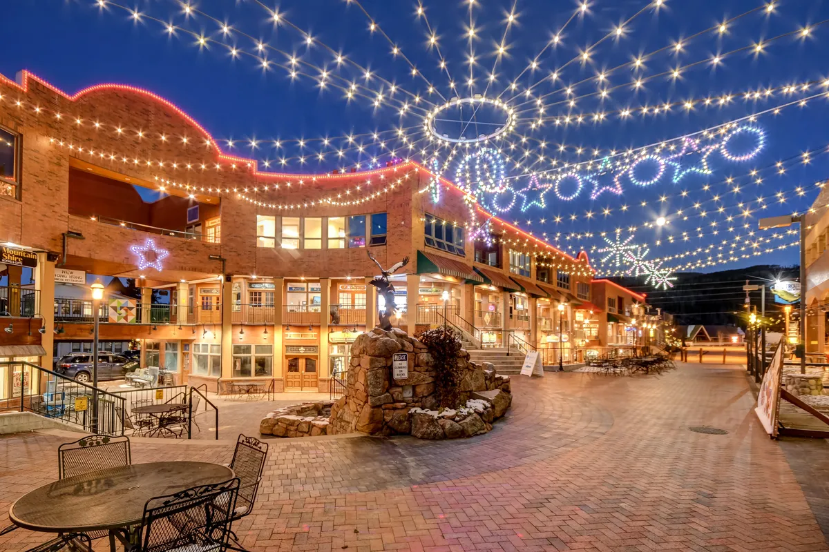 A lively streetscape adorned with twinkling lights, decorative stars, and a stone fountain, framed by outdoor seating areas. The scene exudes a festive, welcoming atmosphere, perfect for outdoor dining, shopping, and gathering during seasonal celebrations or holiday events.