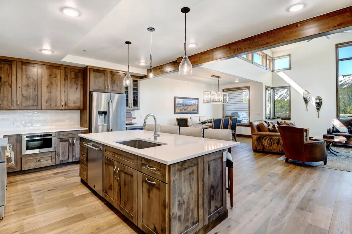 This image features a spacious, inviting kitchen with a prominent island, visible dining area, and adjacent living room seating. The setting is designed for group gatherings, celebrating meals, and culinary activities. The island offers bar seating and is surrounded by high-end appliances, capturing the gourmet atmosphere.